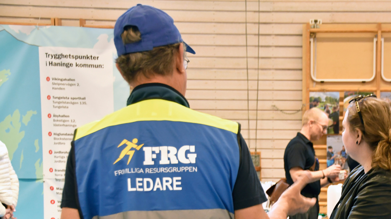 Person med blå keps och väst med texten ”FRG Frivilliga Resursgruppen Ledare” står inomhus vid ett informationsbord. I bakgrunden syns en affisch med rubriken ”Trygghetspunkter i Haninge kommun” och en lista med platser.