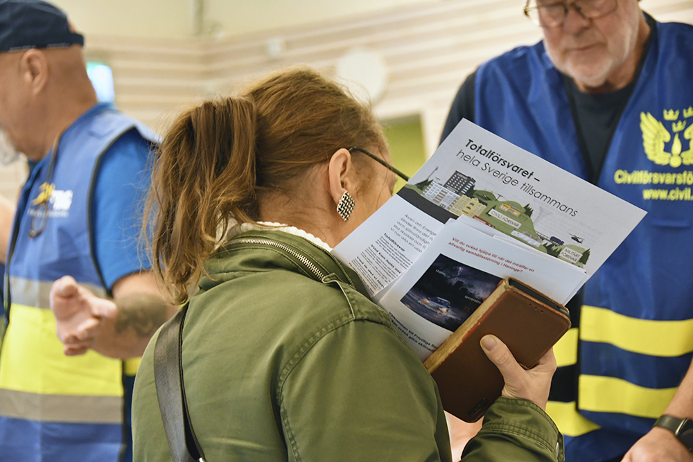 En person läser en broschyr med titeln 'Totalförsvaret – hela Sverige tillsammans', medan två personer i blå västar med civilförsvarstext syns i bakgrunden.