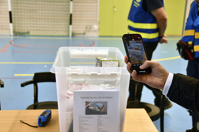 En plastlåda med etiketten 'Vad bör du ha i din krislåda?' visas upp. En person fotograferar innehållet med sin mobiltelefon.