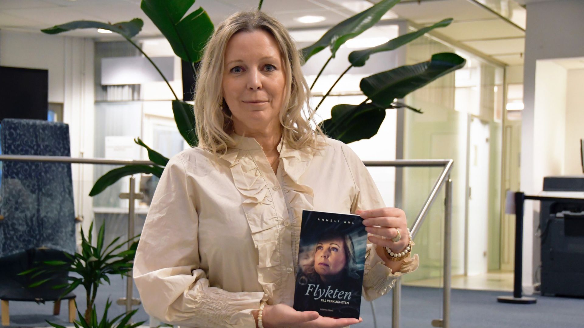 Författaren håller upp sin bok med båda händerna. Boken har en mörk omslagsdesign med titeln "Flykten till verkligheten". Kvinnan bär en ljus, långärmad blus med volanger vid bröstet och ärmarna, samt ett armband med pärlor. Bakgrunden är inomhusmiljö med metallräcken och suddiga detaljer.