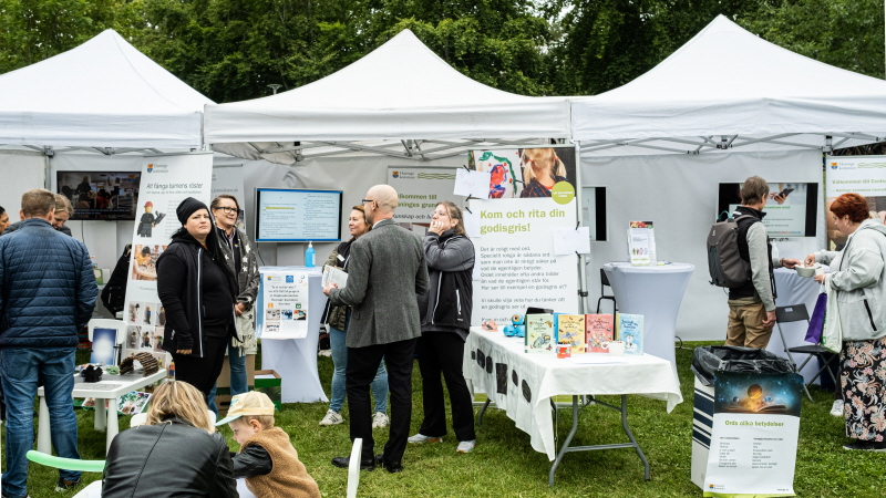 Vita utställningstält på en grön gräsmatta. Vuxna står och samtalar utanför tältet. En liten pojke och mamma sitter vid ett bord och målar.