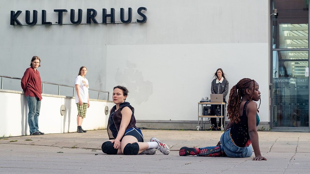 Två personer sitter på marken i förgrunden och tre personer står längre bak, varav en står vid ett bord med teknisk utrustning. 